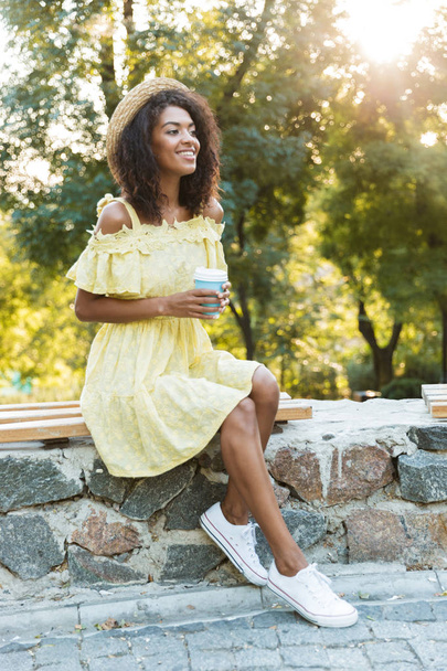 Photo of charming american woman 20s wearing straw hat and dress sitting on bench in park and drinking takeaway coffee - Foto, Imagen