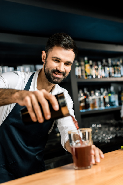 Улыбающийся бармен наливает коктейль из шейкера в стекло за деревянным столиком
 - Фото, изображение
