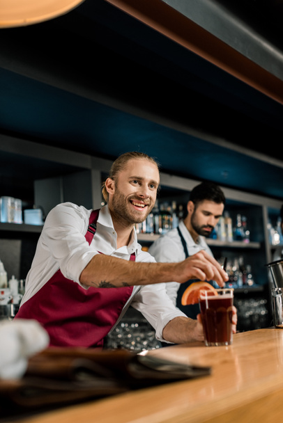 krásný usměvavý barman slouží koktejl na dřevěné počítadlo - Fotografie, Obrázek