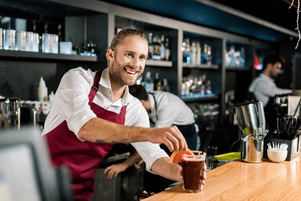 Barman sorridente decorando coquetel com fatia de toranja no balcão de madeira
 - Foto, Imagem