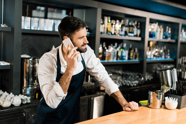 Barman talking on smartphone and standing in apron in bar  - Photo, Image