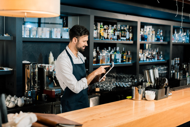Barman standing in apron and typing on digital tablet - Foto, Bild