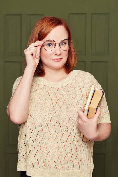 Indoor vertical shot of stupefied European red headed woman keeps hand on rim of spectacles, has unexpected gaze at camera, receives strange news, holds books, posing on classroom door background. - Foto, Imagem
