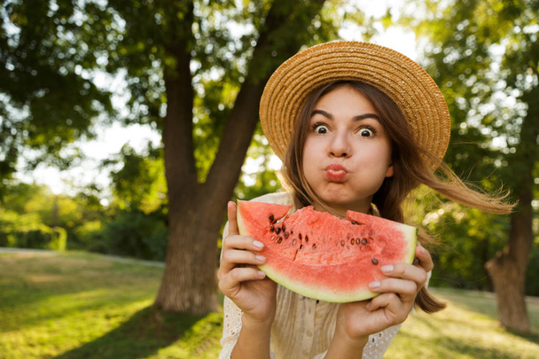 Close up of funny young girl in summer hat spending time at the park, showing slice of a watermelon - 写真・画像