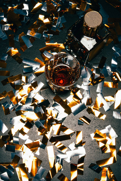 top view of whiskey glass and bottle on table covered by golden confetti  - 写真・画像