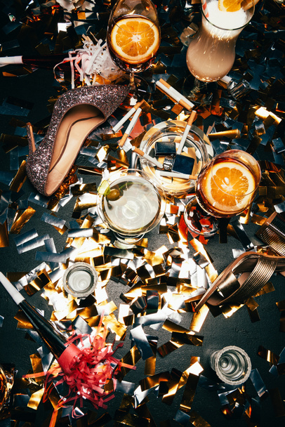 elevated view of female shoes on high heel, ashtray with cigarettes, alcoholic cocktails and party horns on table covered by golden confetti  - Φωτογραφία, εικόνα