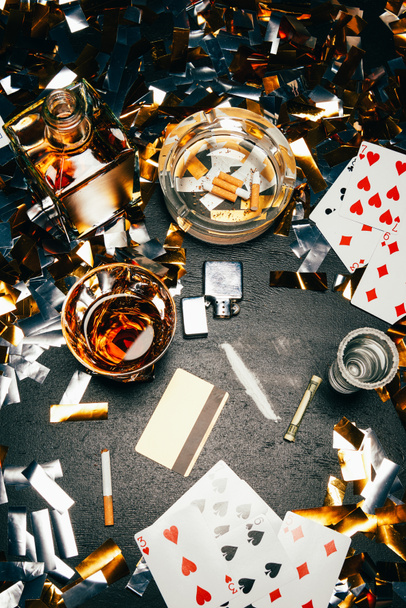 view from above of playing cards, cigarettes, whiskey, rolled banknote, credit card and cocaine on table covered by golden confetti  - Photo, Image