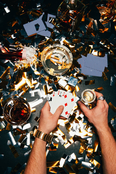 vista parcial del hombre con naipes y alcohol sentado en la mesa cubierto por confeti dorado
 - Foto, Imagen