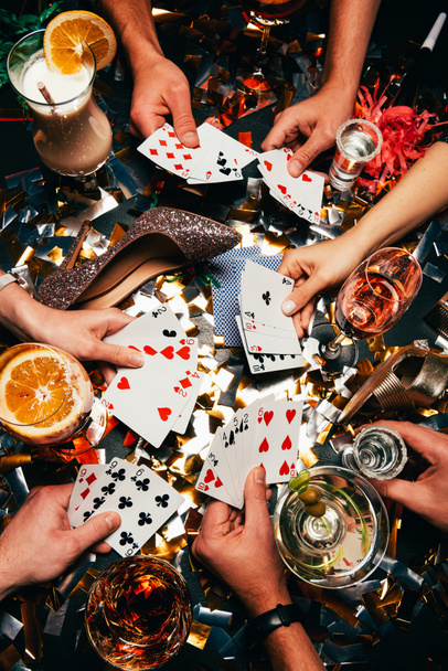 обрезанный снимок друзей с алкогольными коктейлями, играющих в покер за столом, покрытым золотыми конфетти
  - Фото, изображение