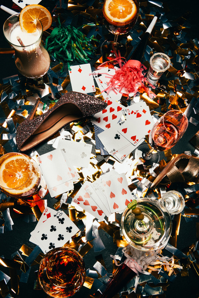 вид женской обуви на высоких каблуках, игральные карты, алкогольные коктейли и рога для вечеринок на столе, покрытом золотыми конфетти
  - Фото, изображение