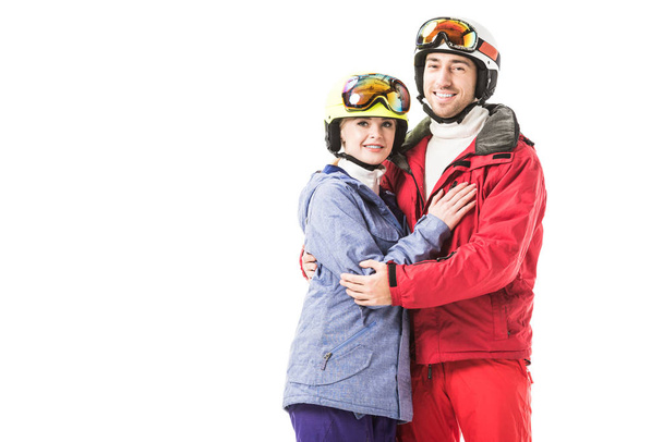 Couple en combinaison de ski, lunettes et casques serrant, souriant et regardant la caméra isolée sur blanc
 - Photo, image