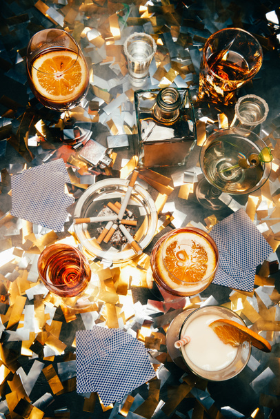 flat lay with cigarettes, alcoholic cocktails, whiskey and playing cards on table covered by golden confetti  - Photo, Image