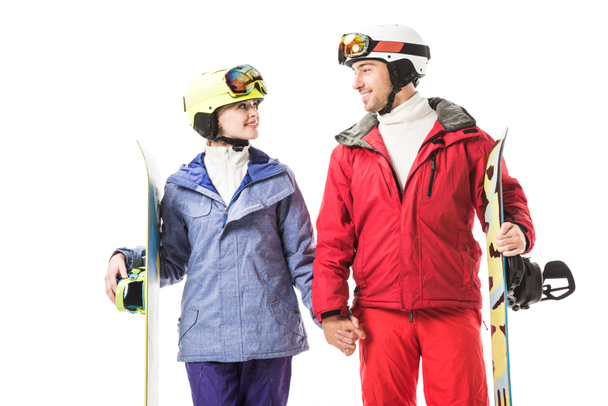 Pareja en ropa de esquí con tablas de snowboard cogidas de la mano, mirándose y sonriendo aislados en blanco
 - Foto, Imagen