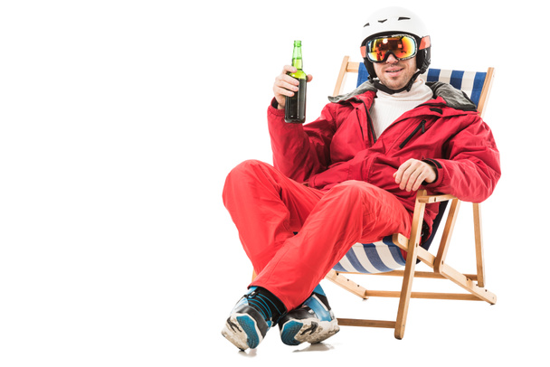 Hombre feliz en traje de esquí rojo con botella de cerveza sentado en la silla de cubierta y sonriendo aislado en blanco
 - Foto, imagen