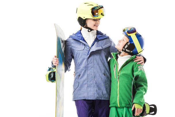 Glimlachend volwassen moeder met snowboard knuffelen preteen zoon in snowsuit geïsoleerd op wit - Foto, afbeelding
