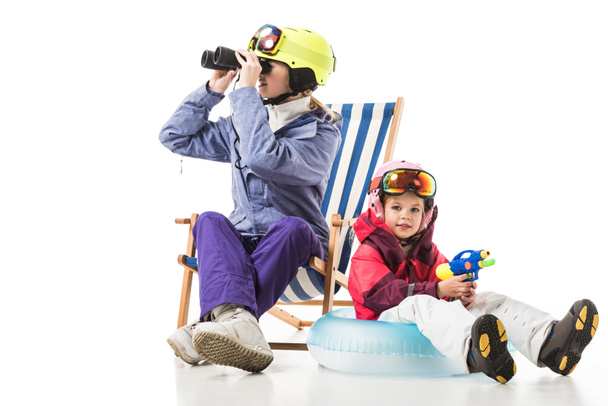 woman with binoculars sitting in deck chair while daughter with water gun sitting on swim ring isolated on white - Photo, image