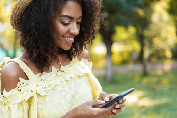Привлекательная молодая африканка в летнем платье прогуливается по городскому парку, держит в руках мобильный телефон
, - Фото, изображение