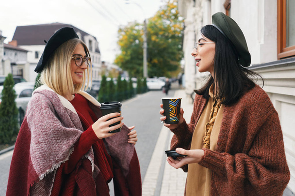 話している 2 つの女性の友人の屋外のポートレート。カジュアルな暖かい服装やメガネの都市の女の子歩く寒い季節と都市背景でコーヒーを飲んでいます。都市生活、友情の概念.  - 写真・画像