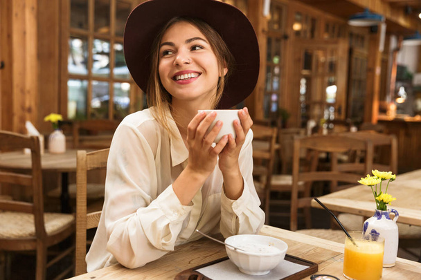 Portrait of smiling woman wearing hat drinking tea while sitting in stylish cafe - Foto, Imagen