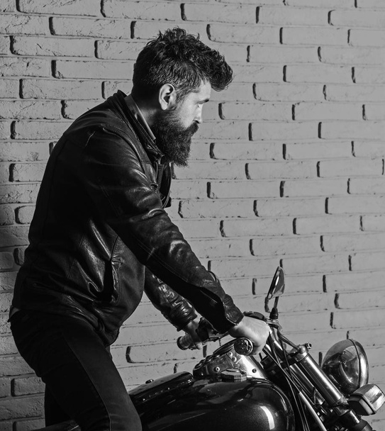 Man with beard, biker in leather jacket near motor bike in garage, brick wall background. Start of journey concept. Hipster, brutal biker on serious face in leather jacket gets on motorcycle - Valokuva, kuva