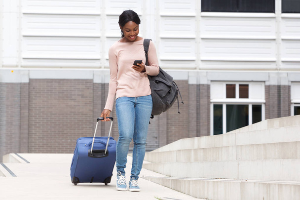 Retrato de cuerpo completo de una joven afroamericana feliz caminando con una maleta y un teléfono celular
 - Foto, Imagen