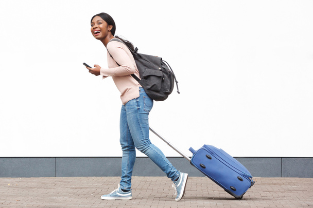 Täyspitkä profiili muotokuva nuori nainen matkustava kävely matkapuhelin ja matkalaukku vastaan valkoinen seinä
 - Valokuva, kuva