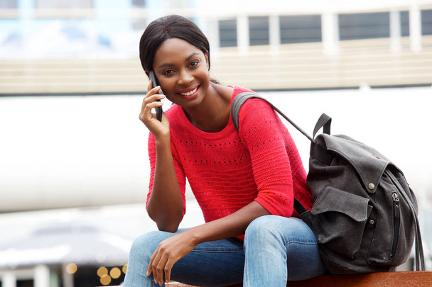 Portrét mladé afroamerické ženy mluví o mobilní telefon zároveň sedět v městě s taškou - Fotografie, Obrázek