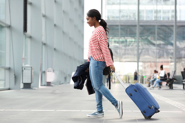 Full length side portrait of young black woman walking with suitcase in airport - 写真・画像