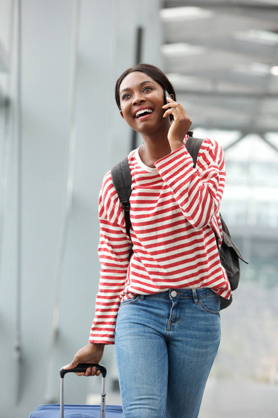 Portrait of happy young african american woman walking and talking with cellphone in airport terminal - Foto, afbeelding