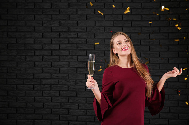 Beautiful young woman with glass of champagne and falling confetti near dark brick wall - Photo, Image