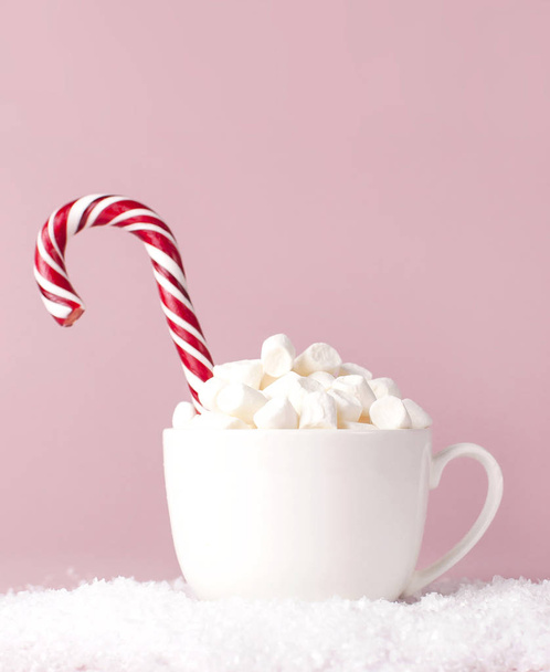 Christmas New Year drink, white mug with marshmallows and Candy Cane in the snow on pink background Flat Lay copy space. Winter traditional drink food. Festive decor celebration Xmas holiday 2019 - Photo, image