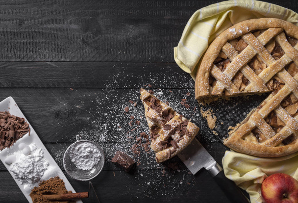 American apple pie and a slice on a knife, with powdered sugar, chocolate, and cinnamon. Above view. On a rustic table. Ready to eat dessert concept. - Photo, Image