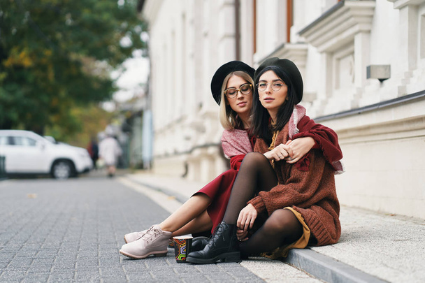 Δύο φίλες πορτραίτο σε εξωτερικούς χώρους. Κορίτσια σε casual ζεστά ρούχα, γυαλιά και μπερέ κάθεται και αγκαλιάζει σε αστικό φόντο, αντιγράψτε χώρο. Τρόπου ζωής, η φιλία και η αδελφότητα έννοια πόλη.  - Φωτογραφία, εικόνα