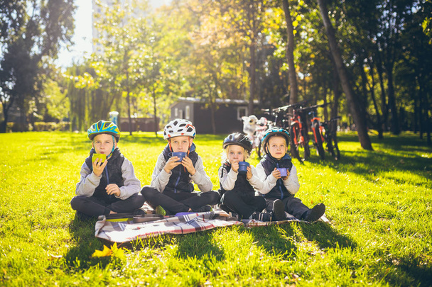 Temat aktywnych wakacji rodzinnych charakter. grupa ludzie małe dzieci troje braci i sióstr siedzieć na kocu w pobliżu rowerów w parku zielona trawa trawnik odpoczynku i pić drinka z filiżanek i herbaty termos. - Zdjęcie, obraz