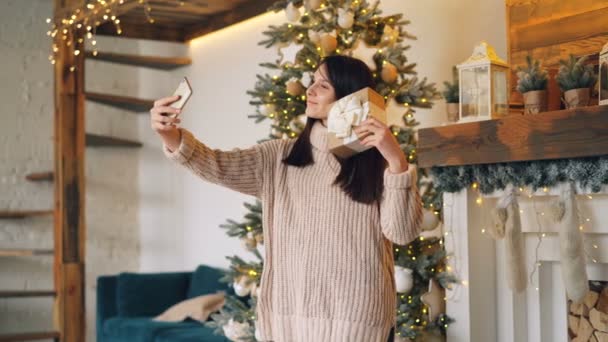 Happy young lady in warm sweater is taking selfie with gift box on Christmas day standing near fireplace and decorated New Year tree. Presents and holidays concept. - Materiał filmowy, wideo