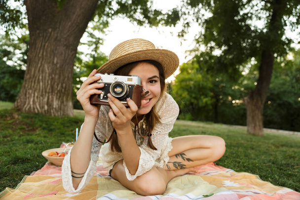 Усміхнена молода дівчина в літньому капелюсі має пікнік у парку, сидить на траві, фотографує фотоапарат
 - Фото, зображення