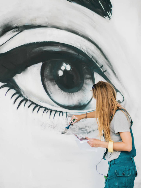Beautiful girl making graffiti of big female face with aerosol spray on urban street wall. Creative art. Talented student in denim overalls drawing picture - 写真・画像