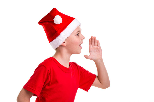Lindo niño sonriente, sombrero de santa en la cabeza, con la mano izquierda verticalmente, llamando. Concepto: Navidad o Feliz Año Nuevo
 - Foto, Imagen