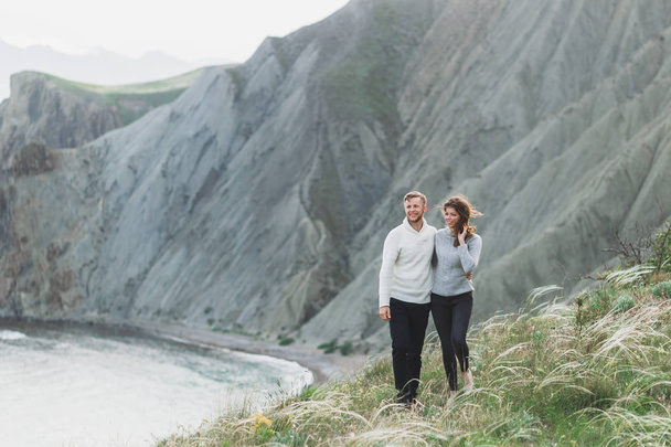 春、カジュアルなスタイルの服セーターとジーンズ マウンテン ビューと北欧の海岸を歩く若いカップル - 写真・画像