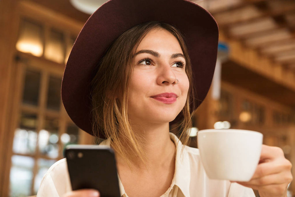 Close up of a lovely girl in hat sitting at the cafe table indoors, holding cup of tea, using mobile phone - Fotografie, Obrázek