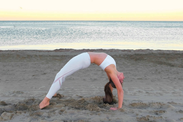 Young woman is doing yoga Dhanurasana Bow Pose on the sand beach at sunrise in autumn. - Фото, зображення