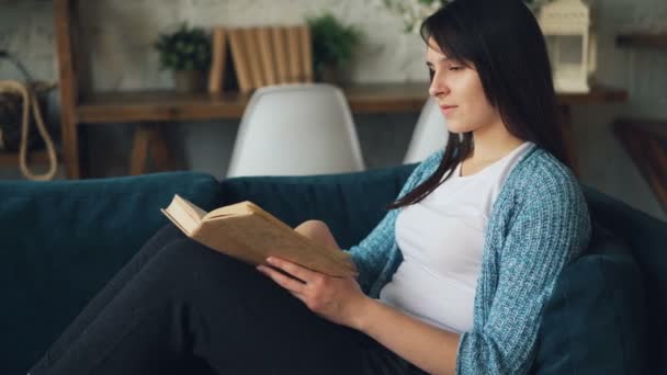 Attractive young lady is reading book and smiling sitting on sofa at home and relaxing alone. Interesting literature, hobby and intelligent people concept. - Felvétel, videó