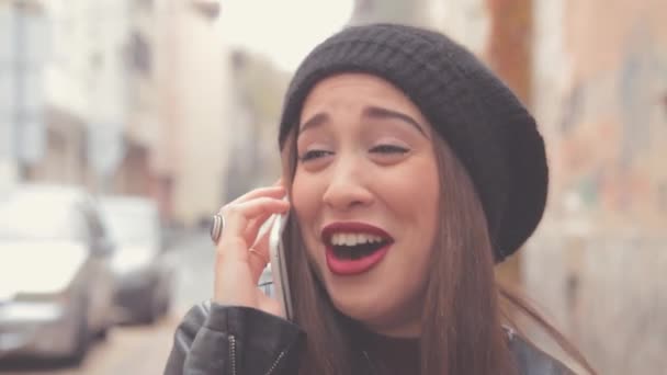 Young brunette woman with red lipstick and beanie hat using smartphone on blurred city background - Footage, Video