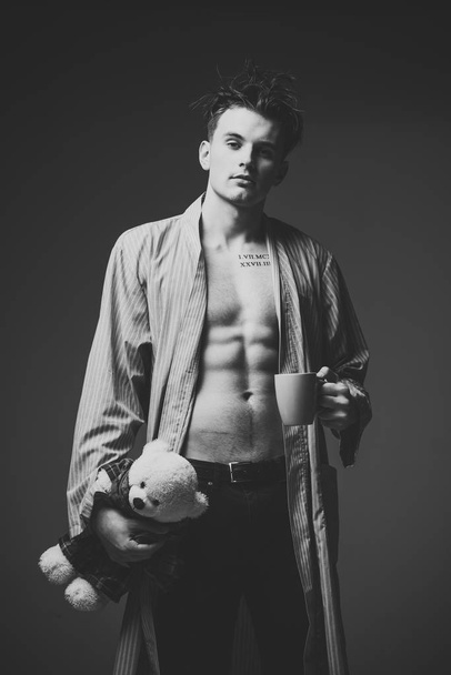 Guy in bathrobe with romantic gift. Man on calm face, muscular figure, holds soft toy plush bear and mug. Romantic morning concept. Man with muscular torso, looks attractive, dark background. - Foto, imagen