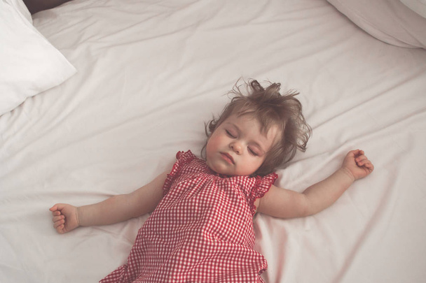 Baby girl sleeping on back with open arms and without pacifier in a bed with white sheets. Peaceful sleeping in a bright room. Pastel retro toned. Soft focus - Foto, imagen