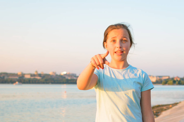 La chica muestra su dedo índice en la dirección de la lente sobre el fondo del estanque y la ciudad
 - Foto, Imagen