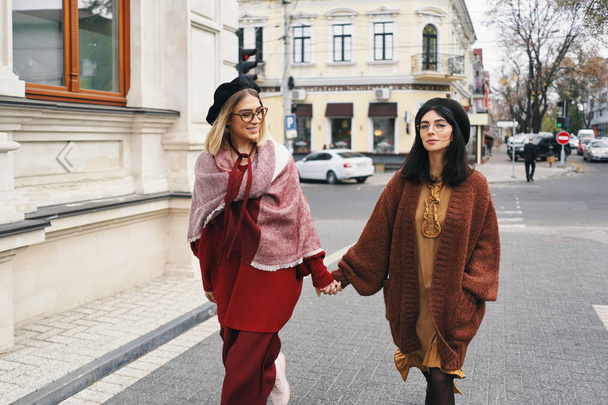 Carefree fashionable women models in elegant autumn clothes and glasses. Young fashion girls in stylish woven wool clothes and hats over urban city background, autumn portrait.  - Foto, imagen