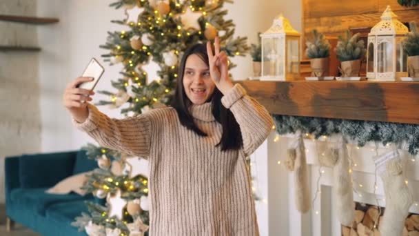 Beautiful young woman is taking selfie with smartphone on Christmas standing near decorated fireplace mantel and New Year tree. Holiday and photo concept. - Filmagem, Vídeo