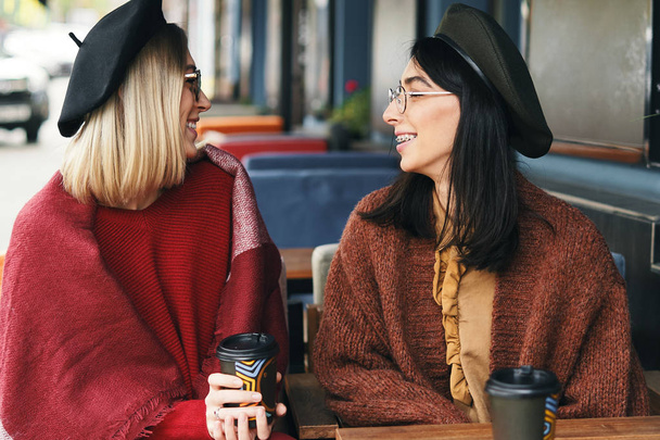 Autumn winter portrait of two young women in an outdoor cafe, drinking coffee to go, talking. Urban background. Sisters - Photo, Image