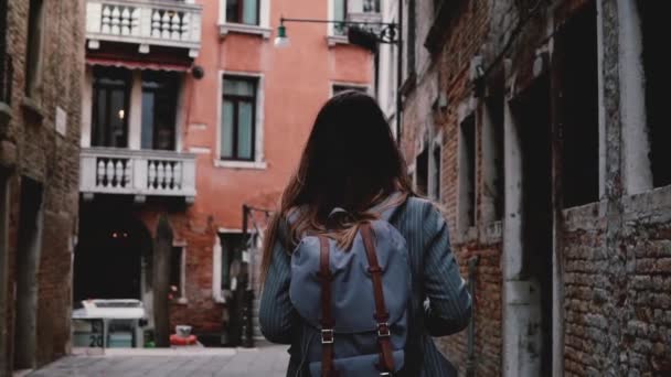 Camera follows young female tourist with backpack walking along beautiful dark ancient city street in Venice slow motion - Video